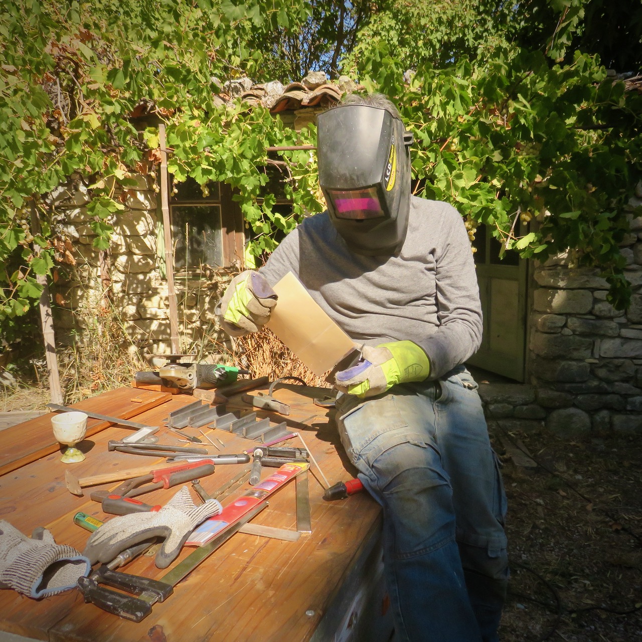 l'Artisan se tiens assis sur son plan de travail encombré et tente de glisser une feuille plastifiée dans un SAMM tout chaud. Il a son casque de soudeur vissé sur la tête, on ne voit pas son visage.